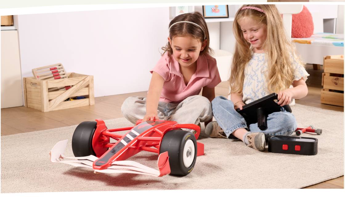 Two girls sitting on a rug playing with a red toy Formula 1 car and its remote control.