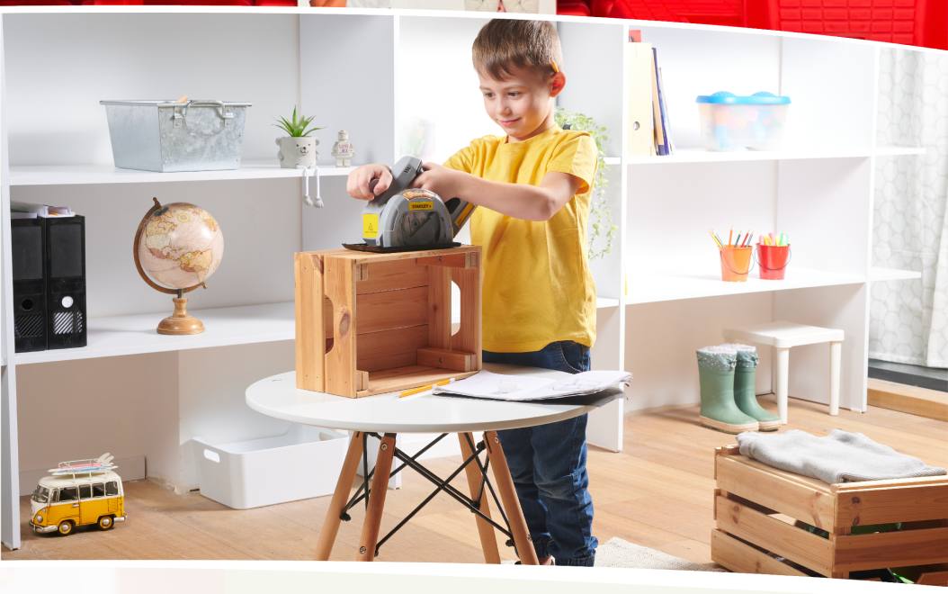 A boy in a yellow T-shirt using a toy circular saw on a wooden crate at a play table.