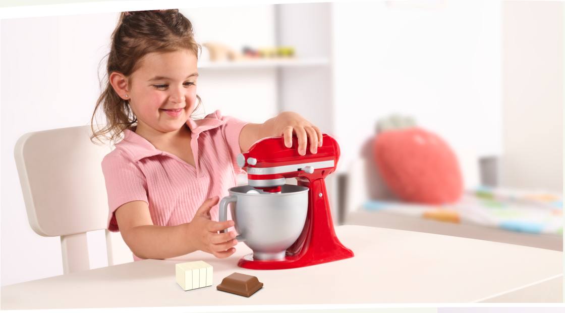 A girl in a pink shirt using a red toy stand mixer at a kitchen counter.